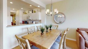 A dining area with a wooden table, chairs, a chandelier, and a kitchen in the background. - Lakefront condo with pool, tennis, lake views, grill, patio, W/D, fast WiFi & AC (Naples)