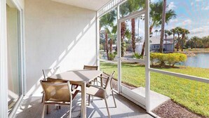 A covered patio with a table and chairs, large windows, and a view of a lake with palm trees. - Lakefront condo with pool, tennis, lake views, grill, patio, W/D, fast WiFi & AC (Naples)