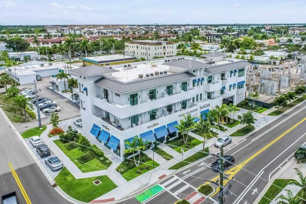 Un edificio de varios pisos con diseño moderno, que destaca por una entrada prominente, un área de estacionamiento y un espacio verde con palmeras.