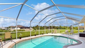 A swimming pool with a covered area, surrounded by a white metal frame, and a grassy area with chairs and a building in the background. - Close to Gulf with pool & updated interior (Rotonda West)