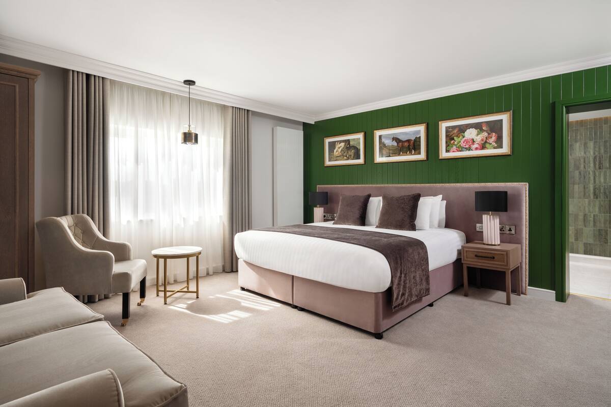 A hotel room with a large bed, a sitting area with a chair, a small table, and a nightstand with a lamp.
