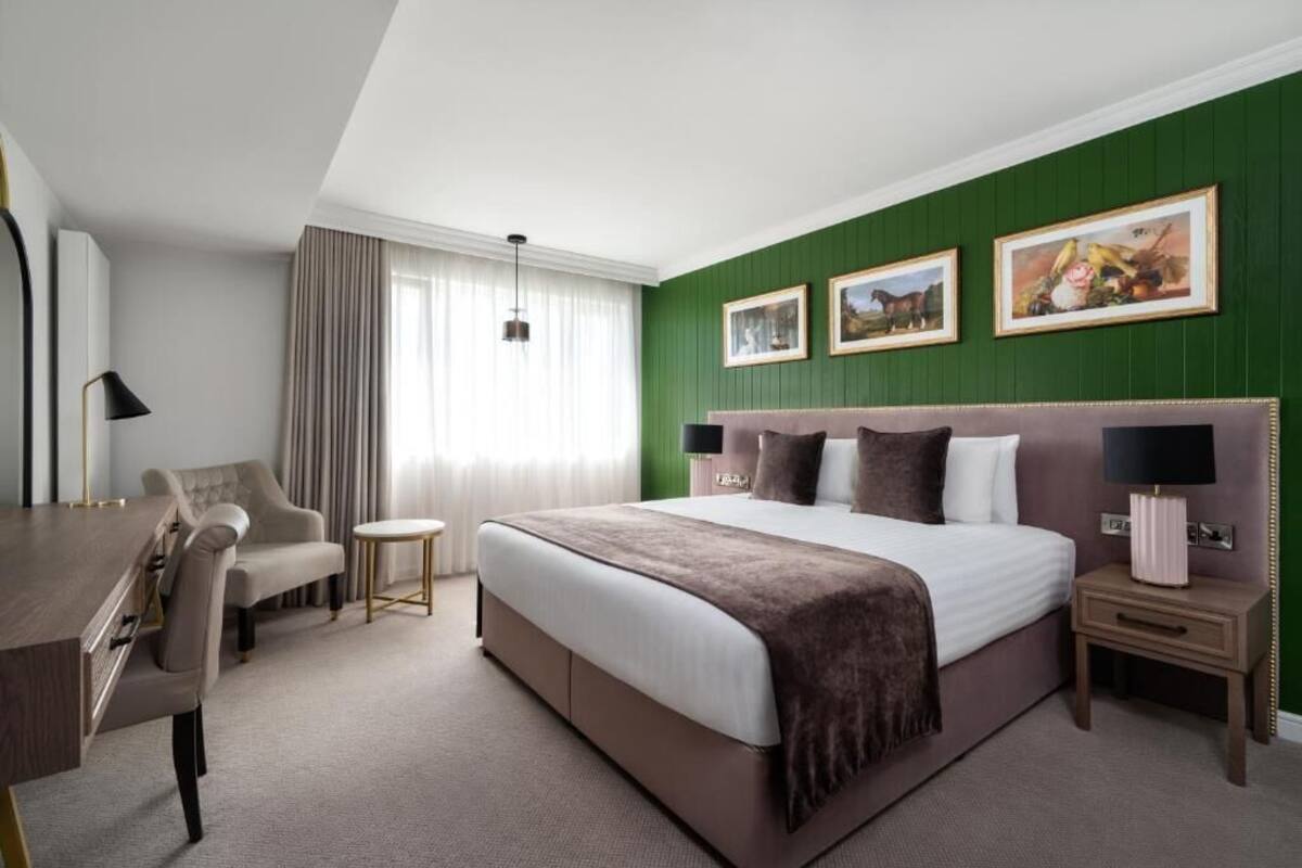 A hotel room with a large bed, a desk, a chair, a small table, and a nightstand.