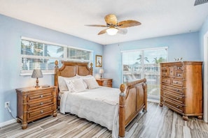 A bedroom with a bed, wooden nightstands, a dresser, a ceiling fan, and windows with blinds.