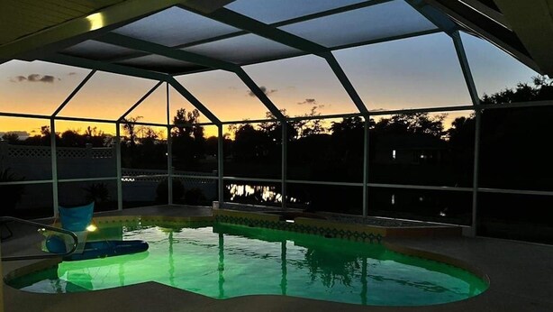 An indoor swimming pool with a glass ceiling, surrounded by a fence, and a view of the sunset. - Bohemian canal-front retreat with pool, beach gear, adult bikes, & AC - dogs ok (Rotonda West)