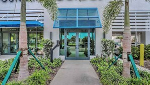 A building entrance with a blue awning, palm trees, and a pathway leading to the door. - Exceptional condo with balcony, WIFi, and located close to beaches and downtown (Naples)