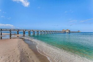Ein Strand mit einer hölzernen Mole, die ins Wasser ragt, einem sandigen Ufer und blauem Himmel.