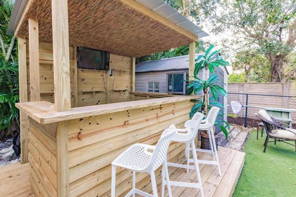 Un espace bar extérieur en bois, doté d’un toit de chaume, d’un ensemble de sièges composé de chaises en plastique blanc, et dont l’arrière-plan est constitué d’une maison et de verdure.