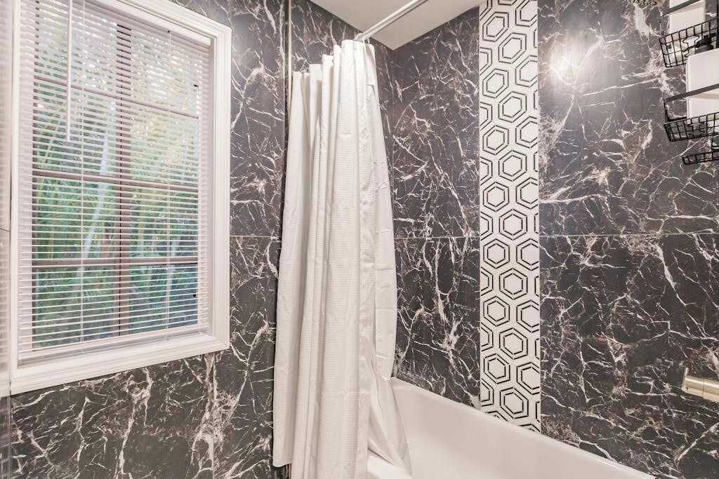 Ein Badezimmer mit Duschvorhang, Fenster mit Jalousien und einer Marmorwand mit geometrischem Muster.