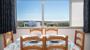 Un coin repas avec une table dressée pour quatre personnes, une vue sur un paysage côtier par la fenêtre et une nappe à motifs.