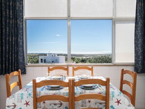 Ein Essbereich mit Tisch für vier Personen, Blick auf eine Küstenlandschaft durch ein Fenster und einer gemusterten Tischdecke.