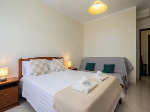 A double bed with a wooden headboard, two bedside tables with lamps, a grey sofa with green cushions, and a beige throw blanket on the bed. - Elymus Apartment, Tavira, Algarve (Tavira)