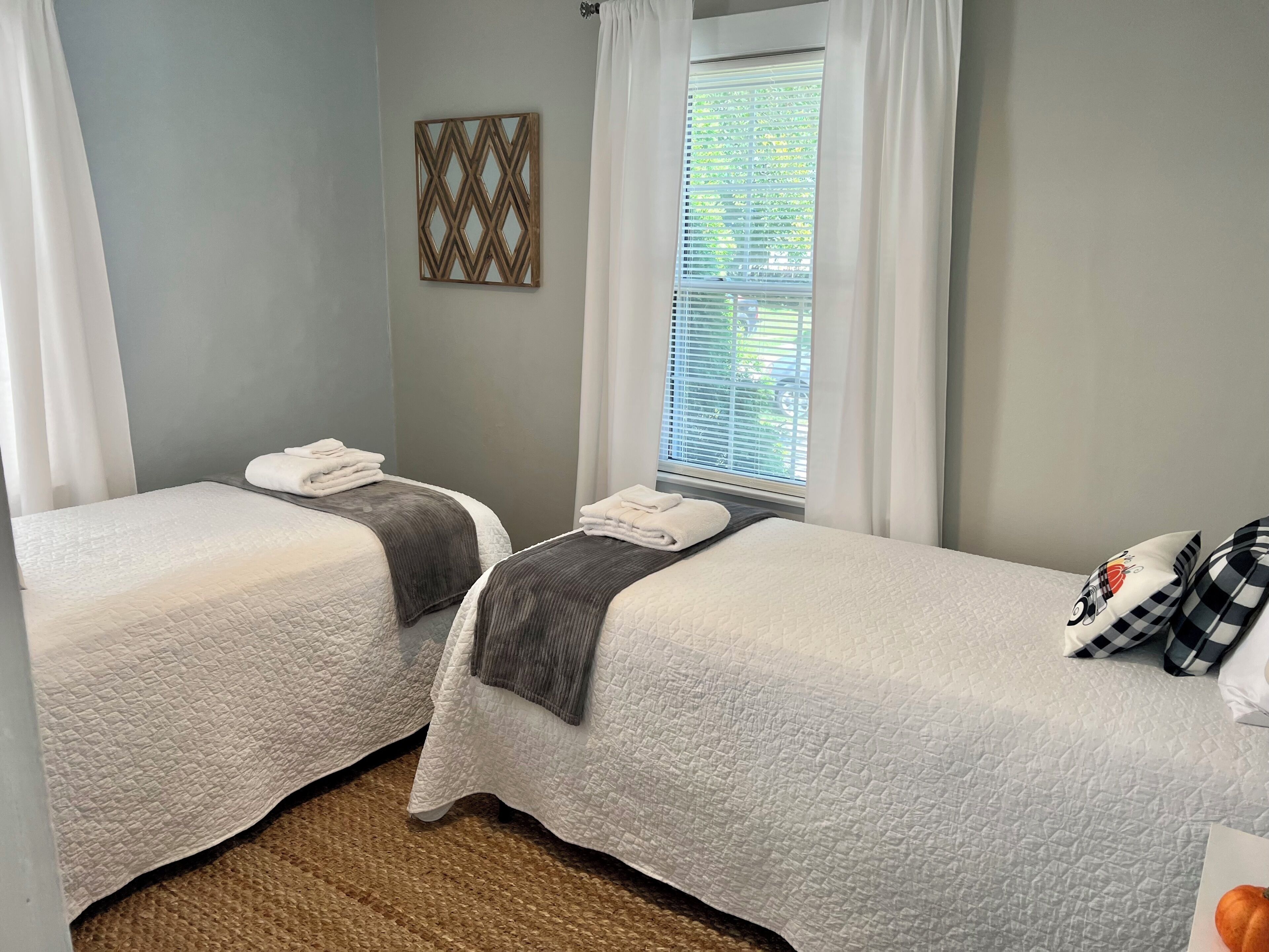 A hotel room with two beds, a window with blinds, a wall-mounted decorative piece, and a woven rug.