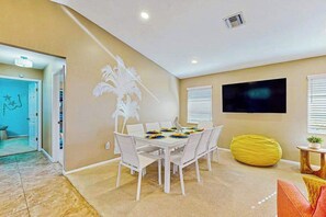 Un comedor con una mesa blanca dispuesta para seis personas, un sillón de pie amarillo y un televisor de pantalla plana en la pared.