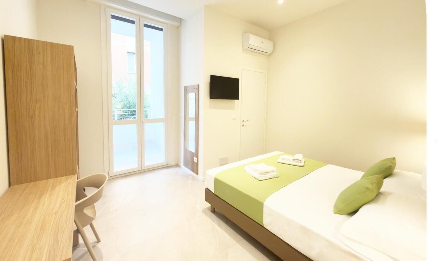 A hotel room with a bed, a desk, a chair, a TV, and a sliding door to the outside.