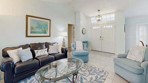 A living room with a black leather sofa, a blue armchair, a glass coffee table, a ceiling fan, and a framed picture on the wall. - Dog-friendly home w/private salt water in ground pool & close to Shamrock Park (Venice)