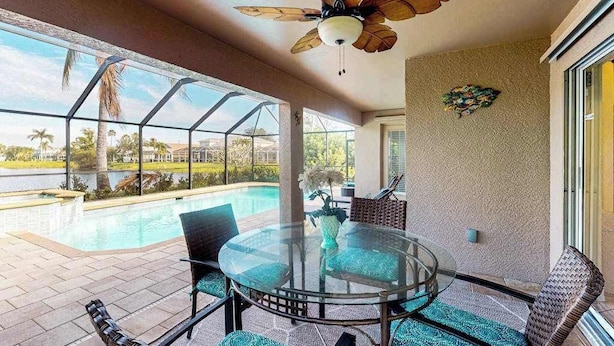A glass-top table with chairs, a ceiling fan, and a view of a swimming pool. - Beautiful Lakefront Home with Private Pool/Spa, Furnished Lanai, Grill, & WiFi (Fort Myers)