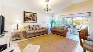 A living room with two patterned sofas, a TV, a painting, and a chandelier. - Beautiful Lakefront Home with Private Pool/Spa, Furnished Lanai, Grill, & WiFi (Fort Myers)