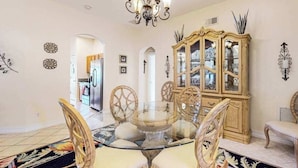 A dining area with a glass-top table, wicker chairs, a chandelier, and a wooden cabinet with glass doors. - Beautiful Lakefront Home with Private Pool/Spa, Furnished Lanai, Grill, & WiFi (Fort Myers)