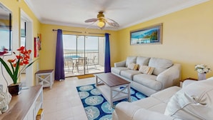 A living room with a sofa, a coffee table, a ceiling fan, a balcony with a view, and a painting on the wall. - Beachfront condo with balcony, community pool, and Wifi (Fort Myers Beach)
