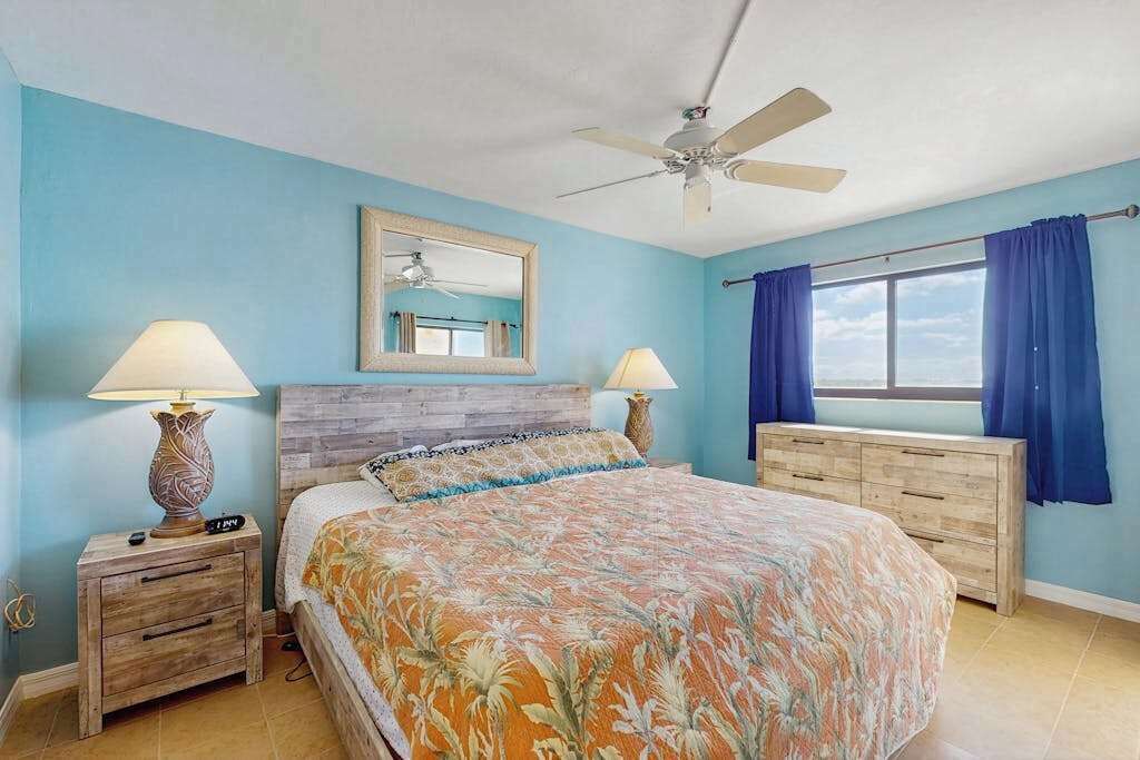 A bedroom with a bed, two nightstands, a ceiling fan, a window with curtains, and a mirror on the wall.