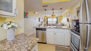 A kitchen with white cabinets, a granite countertop, a dishwasher, a refrigerator, and a window with blue curtains. - Beachfront condo with balcony, community pool, and Wifi (Fort Myers Beach)