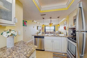 A kitchen with white cabinets, a granite countertop, a dishwasher, a refrigerator, and a window with blue curtains. - Beachfront condo with balcony, community pool, and Wifi (Fort Myers Beach)