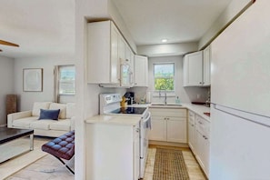 A compact kitchen with white cabinets, a stove, and a sink. There is a brown leather bench and a white sofa in the adjacent living area.