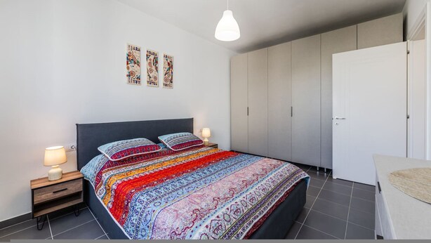 A modern bedroom with a large bed, bedside lamps, a wardrobe, and a hanging light fixture. - Talea Sun Sea Haven 1BR Apartment (Shënkoll)