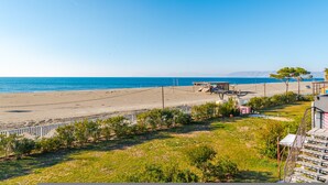 A beach view with a clear sky, a sandy beach, and a few structures including a building with a balcony and a fence. - Talea Sun Sea Haven 1BR Apartment (Shënkoll)
