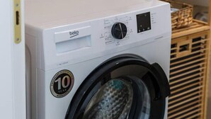 Lavadora de carga frontal blanca con una etiqueta que indica 10 años de garantía, ubicada en una cocina con estantes de madera y piso de baldosas.