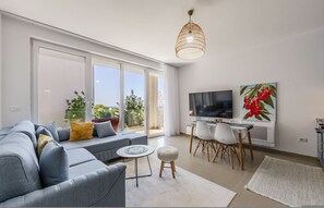 A modern living room with a sofa, a TV, a dining table, and a large window. - Golden Hour Balcony Apartment Pool Beach 100m (Shënkoll)