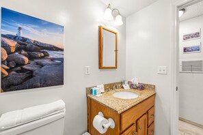 Ein Badezimmer mit Marmorplatte, Waschbecken, Holzschrank, Spiegel und einem Gemälde eines Leuchtturms an der Wand.