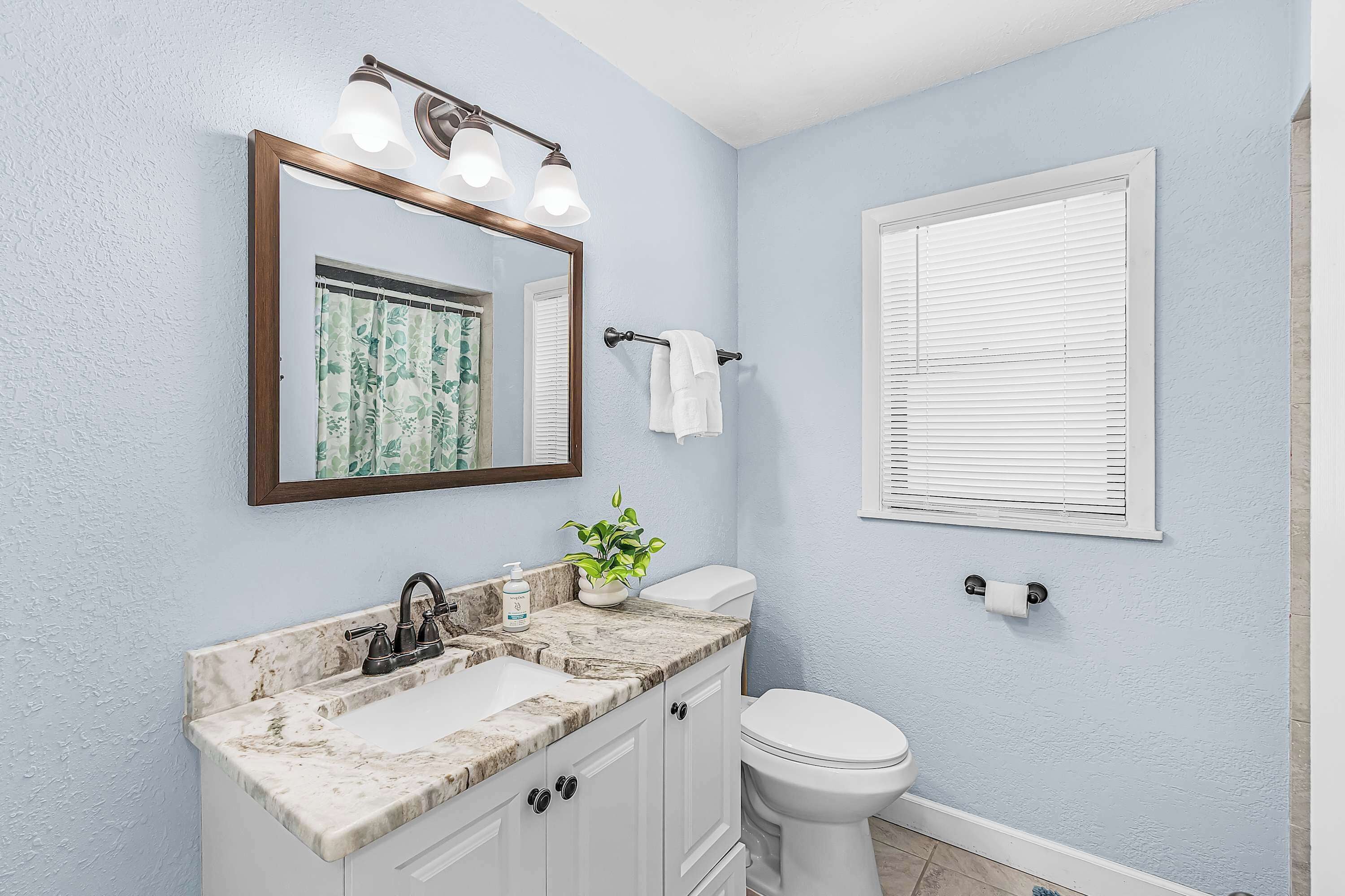 A bathroom with a marble sink, a toilet, a mirror with lights, and a window with blinds.