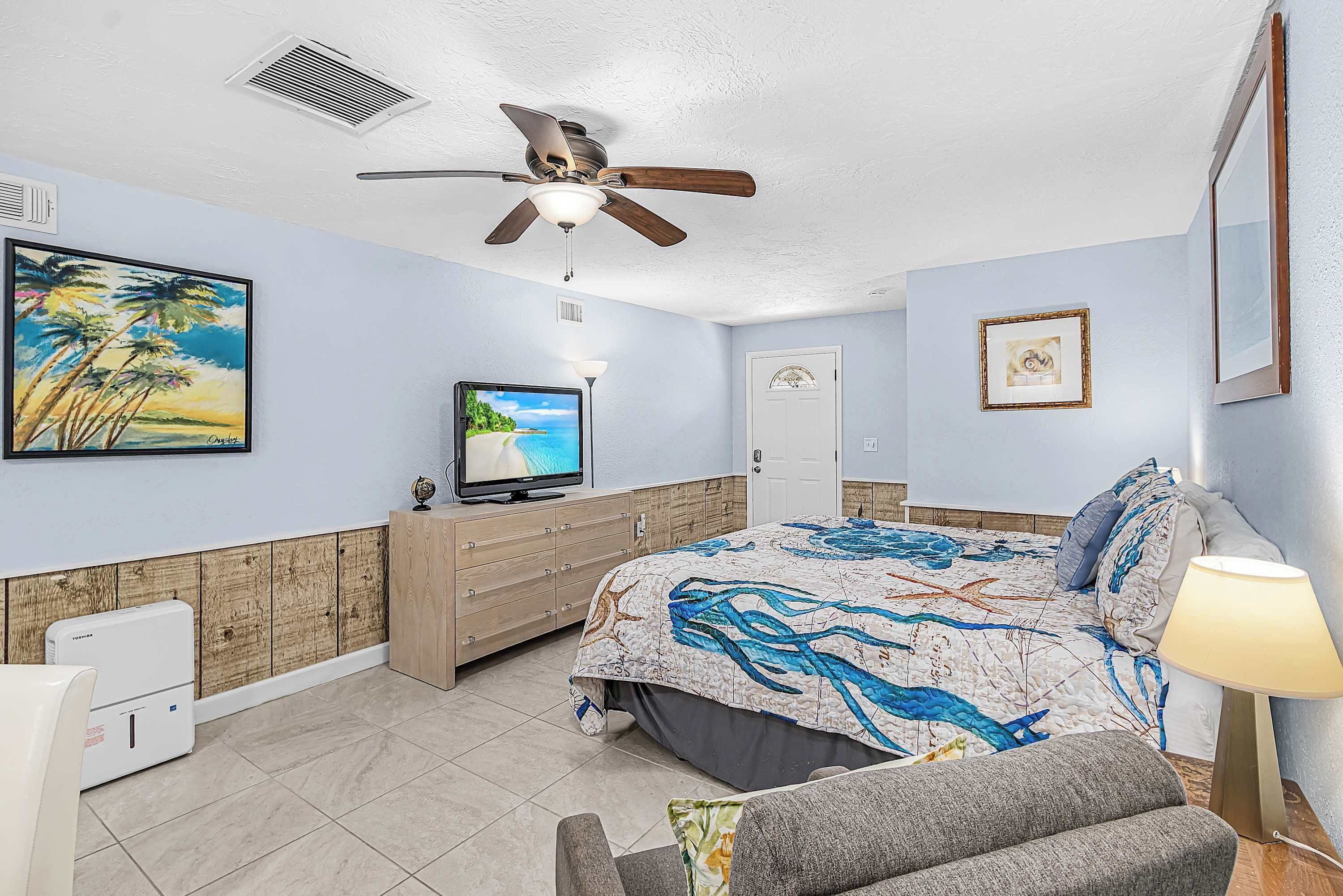 Een slaapkamer met een bed, een televisie, een plafondventilator en een schilderij met een strandthema.
