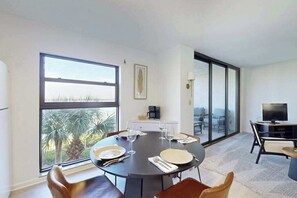 A modern dining area with a round table set for four, large windows with a view of palm trees, a flat-screen TV, and a sliding glass door leading to a balcony. - Spacious condo with water views, tennis, hot tub & pool - ideal for relaxation (Marco Island)
