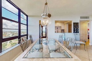 A dining area with a glass table and chairs, large windows, and a view of the city. - Brilliant water view condo w/ shared pool, private washer &dryer, & sunset views (Marco Island)