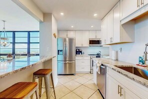 A modern kitchen with white cabinets, stainless steel appliances, and a large window offering a view. - Brilliant water view condo w/ shared pool, private washer &dryer, & sunset views (Marco Island)