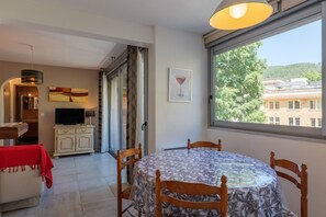 Ein Essbereich mit einem runden Tisch, Stühlen, einem Fernseher auf einem Schrank und einem großen Fenster mit Blick auf Gebäude und Bäume.