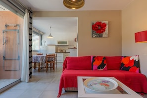 Ein Wohnzimmer mit einem roten Sofa, einem Esstisch mit Stühlen, einer Küche im Hintergrund und einem großen Fenster.