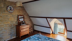 A bedroom with a stone wall, wooden furniture, a bed with a patterned bedspread, a window with curtains, and a sloped ceiling. - Delightful 1-bedroom cottage in tranquil Sainte Price Melrand perfect getaway (Melrand)