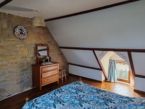 Ein Schlafzimmer mit Steinwand, Holzmöbeln, einem Bett mit gemustertem Überwurf, einem Fenster mit Vorhängen und einer geneigten Decke.