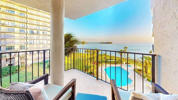 A balcony with wicker furniture, a pool, and a view of the ocean and a building. - Spacious oceanfront condo with heated pool, pickleball, tennis, gym, & balcony (Marco Island)