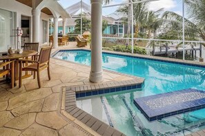 A pool area with a stone patio, wooden furniture, and a view of palm trees. - Luxury canal home with pool, alfresco dining, & walk-in shower - walk to beach (Marco Island)