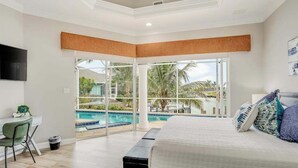 Een slaapkamer met een groot bed, een bureau en een stoel, met uitzicht op een zwembad en palmbomen.