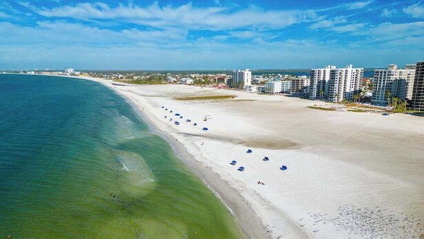 A beach with white sand, clear blue water, and a row of blue umbrellas. - Coastal condo with shared pool, tennis court, and washer & dryer (Fort Myers Beach)