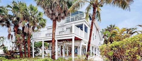 A white two-story house with a balcony, surrounded by palm trees and greenery.