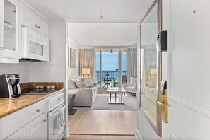 Una habitación de hotel moderna con cocina, un balcón con vistas, una sala de estar con sofá y mesa de centro, y un comedor independiente.