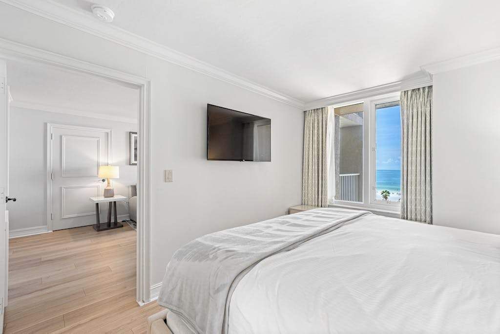 A bedroom with a bed, a television mounted on the wall, a window with curtains, and a view of the ocean.