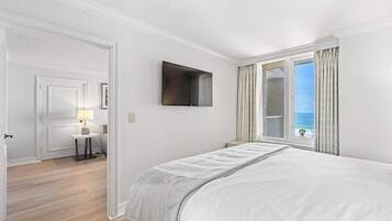 A bedroom with a bed, a television mounted on the wall, a window with curtains, and a view of the ocean.