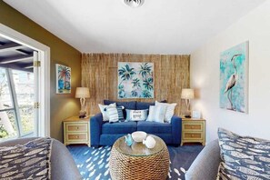 A living room with a blue sofa, two side tables, a glass coffee table, and tropical-themed wall art. - Tropical Palm Island home steps from the beach with grill, outdoor shower, dock (Placida)